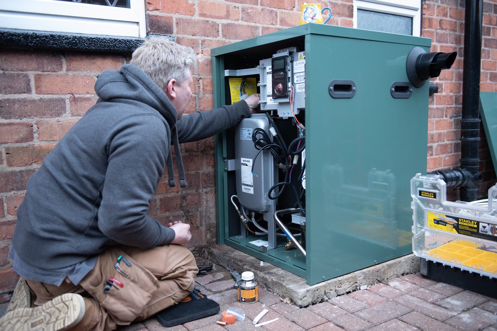James kneeling at an open outdoor boiler with his tool case beside him